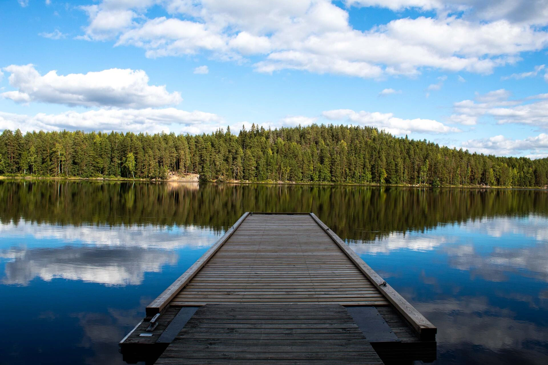 Ponton en bois s’avançant sur un lac calme entouré de forêt, sous un ciel bleu parsemé de nuages